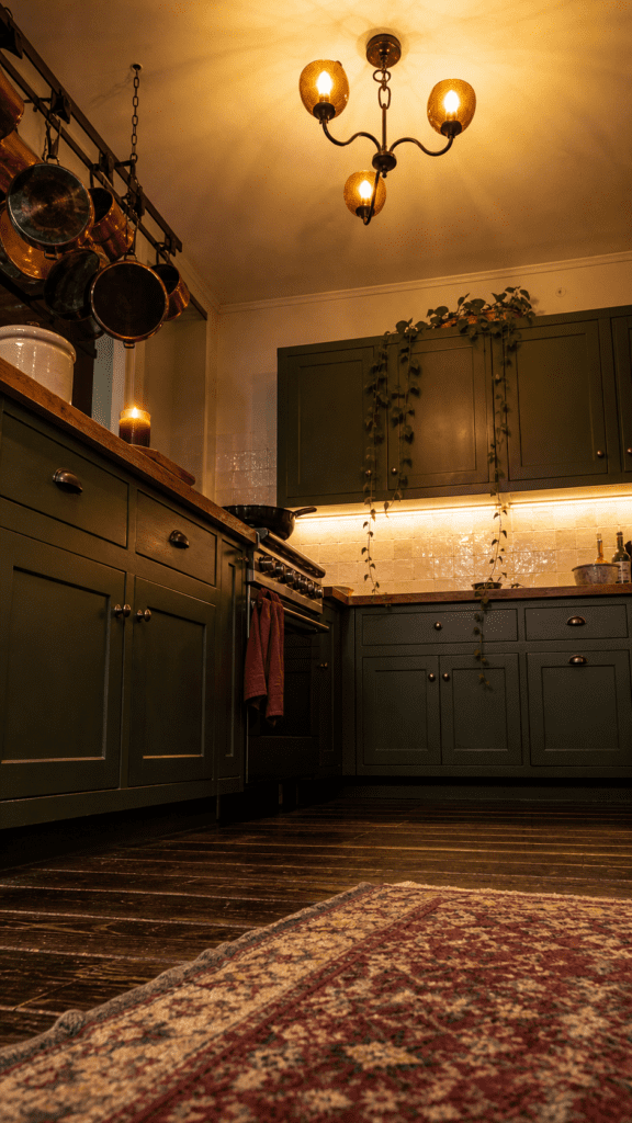 Moody dark-green kitchen at night with a glass globe chandelier, under-cabinet lighting, hanging copper pots, trailing pothos, a lit candle, dark wood floors, and a red Persian rug
