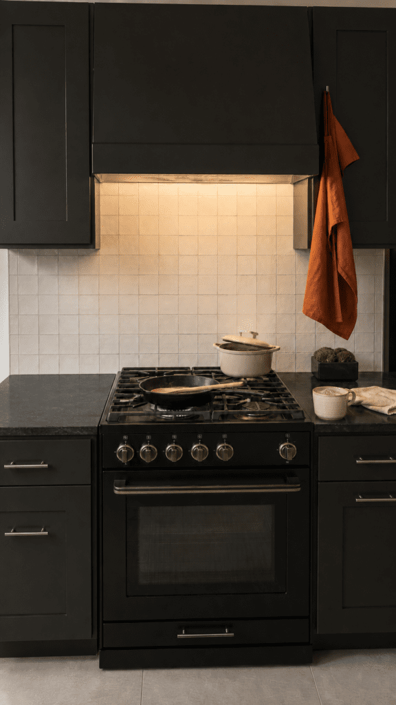 Modern matte-black kitchen with black cabinets and range, soft cream square tile backsplash lit from above, a rust-orange linen towel, cast-iron skillet and Dutch oven on the stove, and a ceramic mug